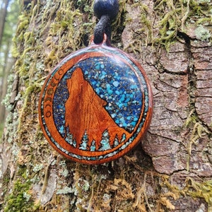 May include: A wooden pendant with a wolf silhouette and a blue and green resin inlay. The pendant is hanging from a black cord.