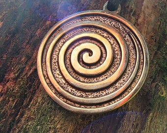 Puede incluir: Un colgante de bronce con un diseño en espiral. El colgante está colgado de un cordón negro.