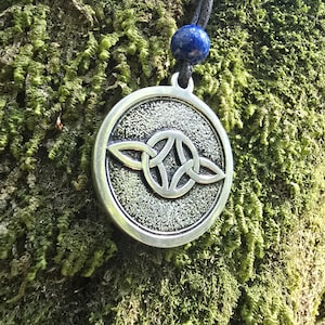Könnte beinhalten: Silberfarbener Anhänger mit keltischem Knotenmuster, an einer schwarzen Kordel mit einer blauen Perle befestigt. Der runde Anhänger hat einen strukturierten Hintergrund. Der Hintergrund ist moosgrün.