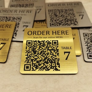 May include: Gold and silver metal table signs with the text "ORDER HERE" and a QR code for online menus. Each sign also displays "TABLE 7". The signs are rectangular with rounded corners.