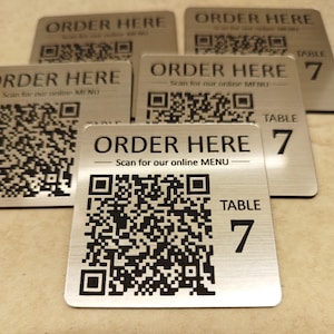 Puede incluir: Señales de mesa de metal cepillado con el texto "ORDER HERE" y un código QR para menús en línea. Cada señal también muestra "TABLE 7". Las señales son cuadradas y tienen un diseño moderno y minimalista.