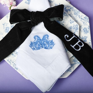 May include: A white linen napkin with blue embroidered initials and a matching design, complemented by a black velvet ribbon. The napkin rests on a blue and white patterned plate, creating an elegant table setting.