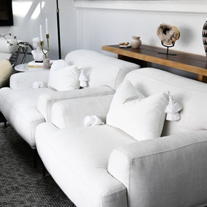 May include: Two white linen armchairs with white tassel pillows, a wooden table with a vase and a decorative stone, and a patterned vase on a wooden shelf.