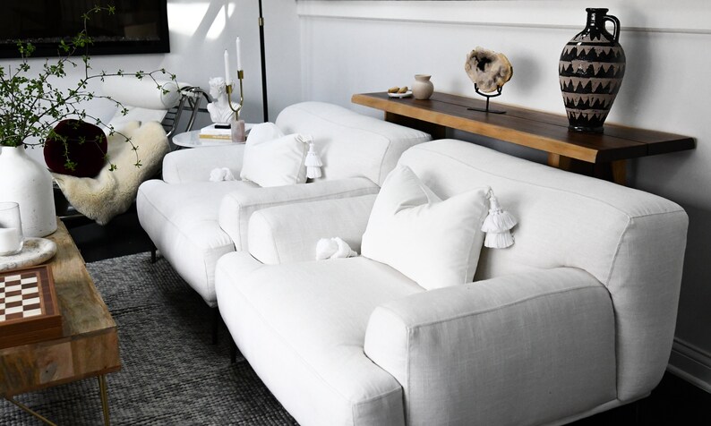 May include: Two white linen armchairs with white tassel pillows, a wooden table with a vase and a decorative stone, and a patterned vase on a wooden shelf.
