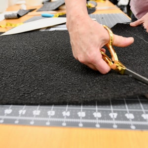 May include: A person's hand uses gold scissors to cut a black fabric on a cutting mat.