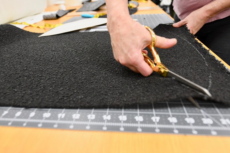 May include: A person's hand uses gold scissors to cut a black fabric on a cutting mat.