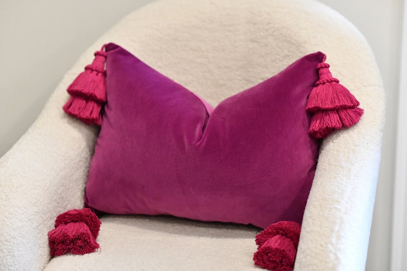 May include: A magenta velvet pillow with tiered tassels at each end rests on a white, textured chair. The pillow's rectangular shape and rich color create a striking contrast against the neutral background, adding a touch of luxury to the decor.
