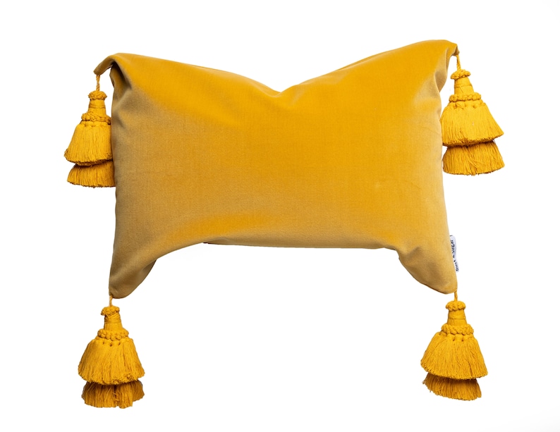 May include: A mustard yellow velvet pillow with four large tassels. The tassels are made of the same fabric as the pillow and are attached to the corners.