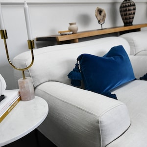 May include: A white couch with a blue velvet pillow with tassels. The couch is in a living room with a white marble side table and a white bust of a woman.