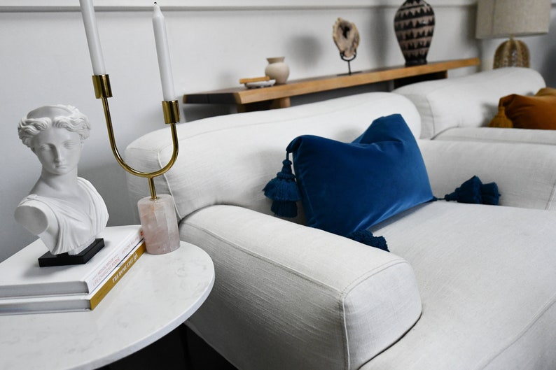 May include: A white couch with a blue velvet pillow with tassels. The couch is in a living room with a white marble side table and a white bust of a woman.