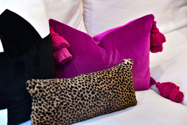 May include: A collection of decorative pillows in various styles and colors. A vibrant magenta velvet pillow with matching tassels is paired with a leopard print pillow and a black pillow. The pillows are arranged on a white surface.