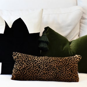 May include: Three decorative throw pillows on a white couch. The pillows are a black and white geometric design, a green velvet pillow with tassels, and a leopard print pillow.