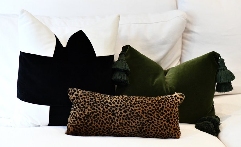 May include: Three decorative throw pillows on a white couch. The pillows are a black and white geometric design, a green velvet pillow with tassels, and a leopard print pillow.