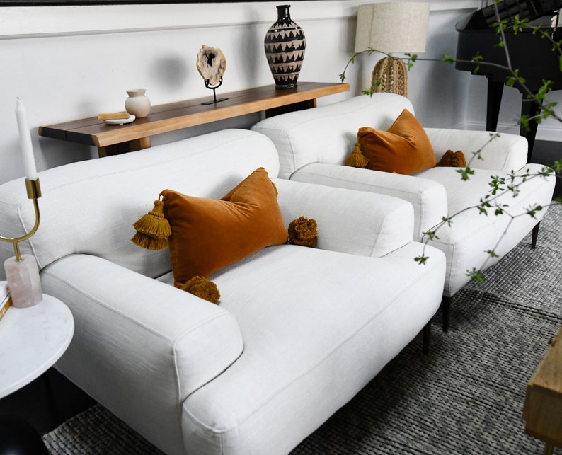May include: Two white linen armchairs with black legs, each with a large orange velvet pillow with tassels and smaller pom-pom accents. The chairs are in a living room setting with a wooden table and a vase.