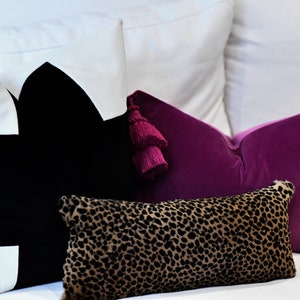 May include: A collection of decorative pillows in various designs and colors. A large black pillow with white accents is next to a vibrant purple velvet pillow with matching tassels. A smaller pillow with a leopard print pattern is in the foreground.