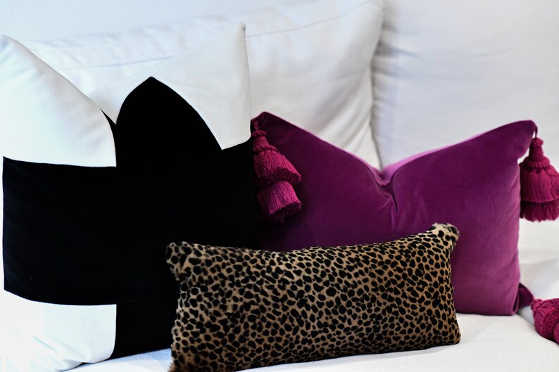 May include: A collection of decorative pillows in various designs and colors. A large black pillow with white accents is next to a vibrant purple velvet pillow with matching tassels. A smaller pillow with a leopard print pattern is in the foreground.