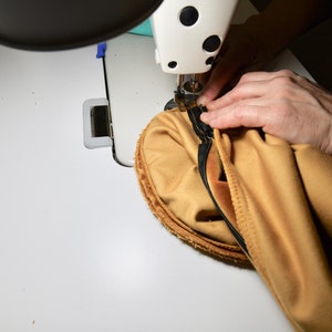 May include: A close-up of a sewing machine needle stitching a seam on a piece of tan fabric. The fabric is being held in place by a hand.
