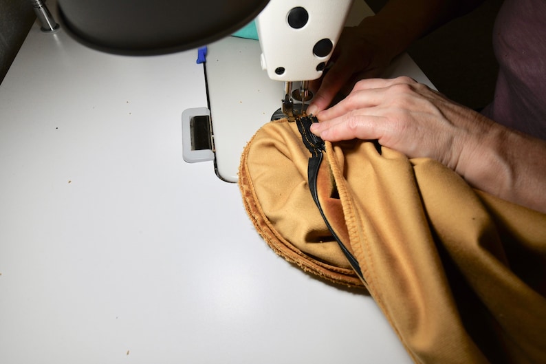 May include: A close-up of a sewing machine needle stitching a seam on a piece of tan fabric. The fabric is being held in place by a hand.