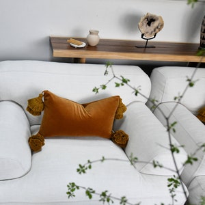 May include: Two white armchairs with plush cushions and throw pillows. The pillows are a deep orange velvet with tassels. The armchairs are in a living room setting with a wooden shelf and a vase on the shelf.