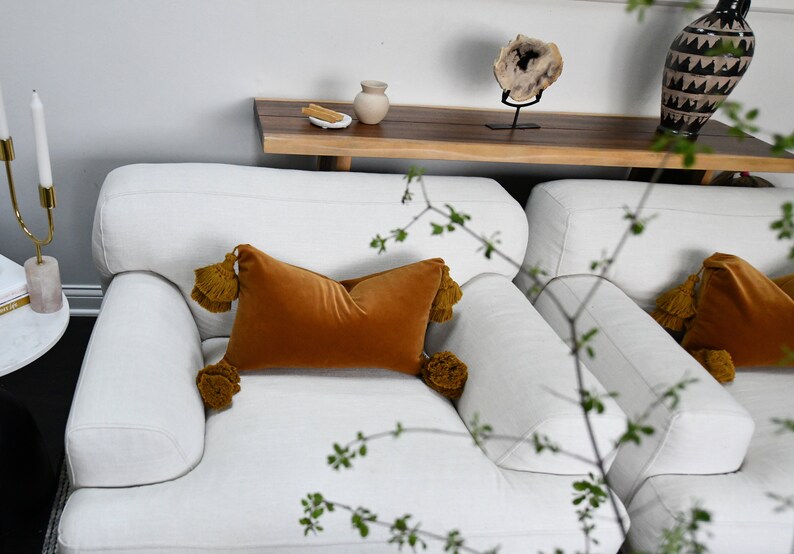 May include: Two white armchairs with plush cushions and throw pillows. The pillows are a deep orange velvet with tassels. The armchairs are in a living room setting with a wooden shelf and a vase on the shelf.