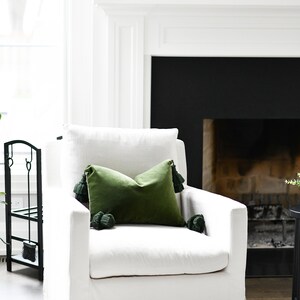 May include: A white swivel chair with a green velvet pillow with tassels. The chair is in front of a fireplace with a black surround.