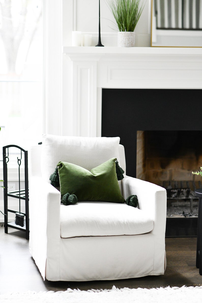 May include: A white swivel chair with a green velvet pillow with tassels. The chair is in front of a fireplace with a black surround.