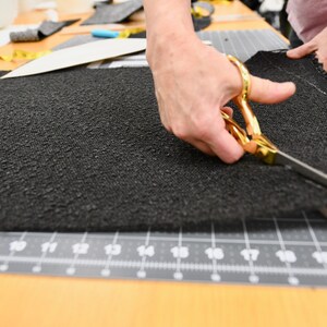 May include: A person using gold scissors to cut a piece of black fabric on a gray cutting mat. The fabric has a textured surface. The scissors are open and positioned to cut along a curved line.