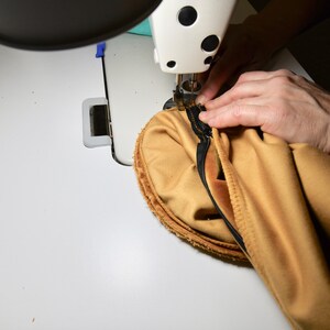 May include: A close-up of a sewing machine needle stitching a seam on a piece of tan fabric. The fabric is being held in place by a hand.