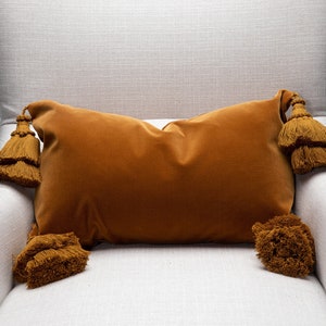 May include: A mustard yellow velvet pillow with large tassels on each corner. The pillow is sitting on a white armchair.