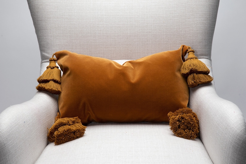 May include: A mustard yellow velvet pillow with large tassels on each corner. The pillow is sitting on a white armchair.