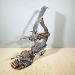 May include: A silver metal sculpture of a high-heeled shoe with a motor and gears attached to it. The shoe is standing on a wooden surface.