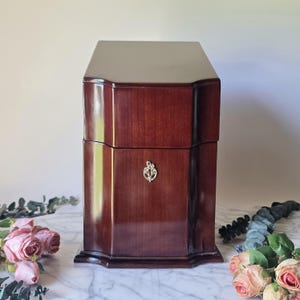 May include: A tall, dark mahogany-colored wooden box with a smooth, polished finish. The box features a tiered, angular lid and a small, silver-toned metallic emblem centered on the front panel. The structure has a rectangular base with beveled edges.