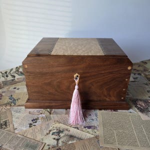 May include: A rectangular wooden box crafted from dark brown wood with a lighter, textured burl wood inlay on the top lid. The box features a small, light-coloured wooden escutcheon at the centre front, from which hangs a soft, pale pink tassel.