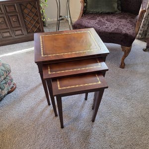 May include: A set of three nested tables made of dark brown wood. Each rectangular table features a smooth top surface with a decorative gold-colored border inlay. The tables are arranged in a cascading stack, with each table decreasing in size.