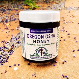 May include: A glass jar of Oregon Osha Honey, labeled "Delicious Herbal Honey" and "Soothing to the throat & uplifting to the soul." The jar is sitting on a sandy surface with purple flower petals scattered around it.