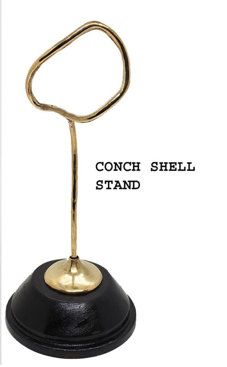 Puede incluir: Un soporte de metal dorado para concha de caracol con una base de madera negra. El soporte tiene una parte superior curva para sostener una concha de caracol.