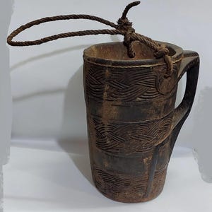 May include: A dark brown wooden cup with a carved design and a rope handle. The cup is made of wood and has a rustic appearance.