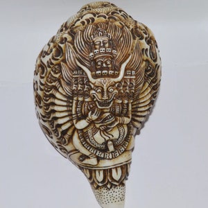 Może przedstawiać: Biała muszla conchowa z misternymi rzeźbami przedstawiającymi wieloheaddowe bóstwo z wieloma ramionami. Bóstwo ma groźny wyraz twarzy i jest ozdobione misternymi klejnotami. Muszla jest tradycyjnym religijnym artefaktem.