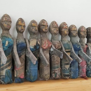 Puede incluir: Una fila de diez muñecas de madera, cada una con una cara pintada diferente y ropa colorida. Las muñecas están dispuestas en línea, con la muñeca más alta a la izquierda y la más baja a la derecha. Las muñecas llevan todas colores y patrones diferentes, y algunas tienen detalles dorados.