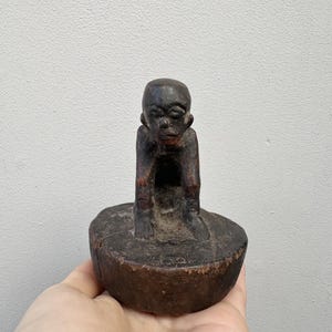 Op de afbeelding: Een donkerbruine houten sculptuur van een zittende figuur met armen die op de knieën rusten. De figuur staat op een ronde, platte basis. De sculptuur heeft een verweerde uitstraling, wat duidt op leeftijd en gebruik. De gelaatstrekken van de figuur zijn zichtbaar.
