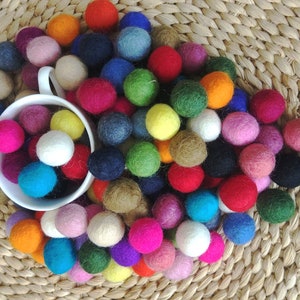 Puede incluir: Una taza blanca llena de bolas de fieltro de colores. La taza está rodeada de más bolas de fieltro de varios colores, incluyendo rojo, azul, verde, amarillo, rosa y morado. Las bolas de fieltro están dispuestas sobre una superficie de mimbre tejido.
