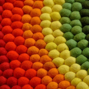 Op de afbeelding: Een kleurrijk rond tapijt gemaakt van viltballen in tinten rood, oranje, geel, groen en bruin. De ballen zijn in een cirkelvormige patroon gerangschikt, waardoor een regenboog-effect ontstaat.