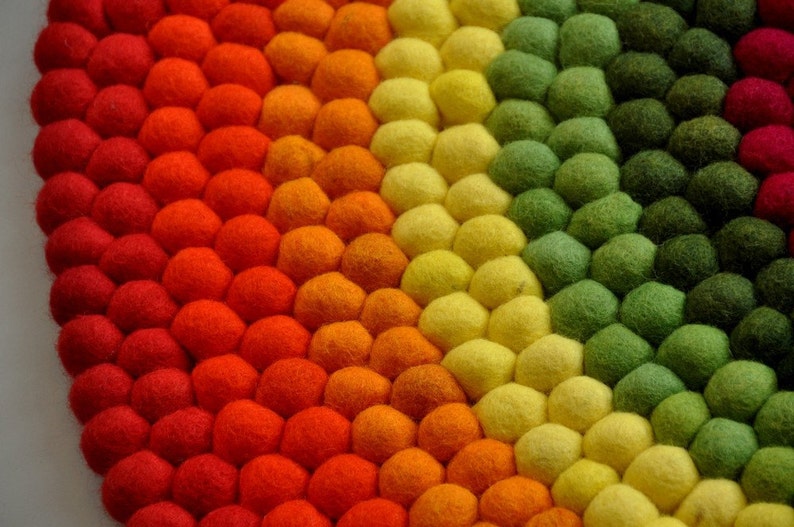 Op de afbeelding: Een kleurrijk rond tapijt gemaakt van viltballen in tinten rood, oranje, geel, groen en bruin. De ballen zijn in een cirkelvormige patroon gerangschikt, waardoor een regenboog-effect ontstaat.