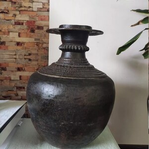 May include: A large, dark brown, ceramic vase with a wide, round body and a narrow neck. The vase has a decorative rim and intricate carvings around the neck.