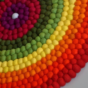 Op de afbeelding: Een kleurrijk rond tapijt gemaakt van viltballen in tinten rood, oranje, geel, groen en paars. De ballen zijn gerangschikt in concentrische cirkels, waardoor een regenboog-effect ontstaat.