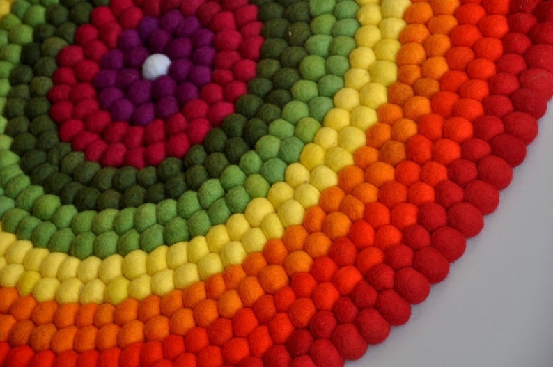 Op de afbeelding: Een kleurrijk rond tapijt gemaakt van viltballen in tinten rood, oranje, geel, groen en paars. De ballen zijn gerangschikt in concentrische cirkels, waardoor een regenboog-effect ontstaat.