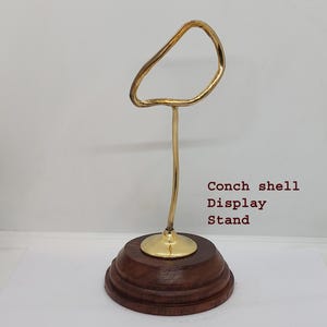May include: A gold metal conch shell display stand with a wooden base. The stand has a curved, open top to hold a conch shell.