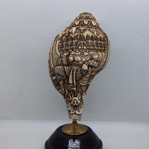 May include: A white conch shell with intricate carvings of a deity with multiple heads and a golden stand. The shell is on a black base.