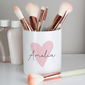 May include: A white ceramic makeup brush holder with a pink heart and the name "Amelia" in black script. The holder contains several makeup brushes with white bristles and rose gold handles. Two bottles are visible in the background.