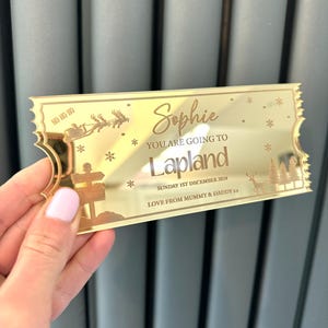 Pode incluir: Bilhete dourado com uma rena e Papai Noel voando no c&eacute;u. O bilhete diz "Sophie You Are Going To Lapland Sunday 1st December 2024 Love From Mummy & Daddy"